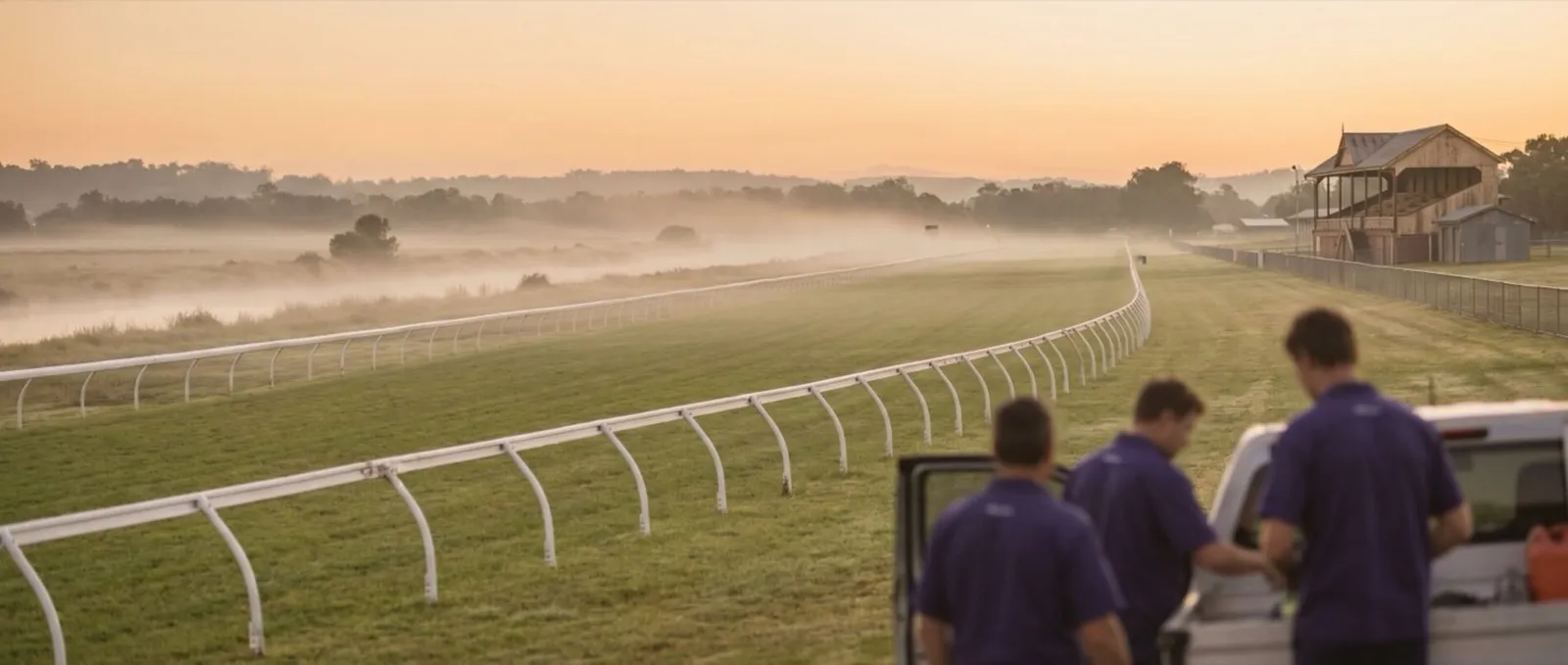 The white running rail and grass track of Wyong Racecourse at dawn, mist over the river flats